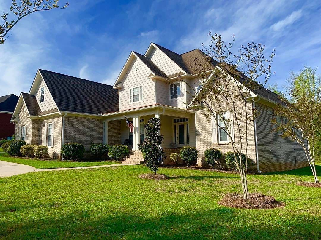 Two-story house with beige siding, a dark roof, and a green lawn under a partly cloudy sky.