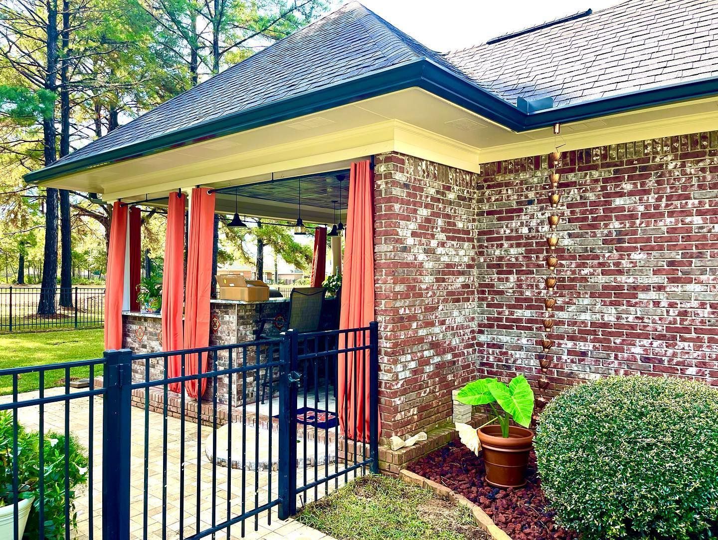 Brick house exterior with covered patio, red curtains, black fence, and potted plants.