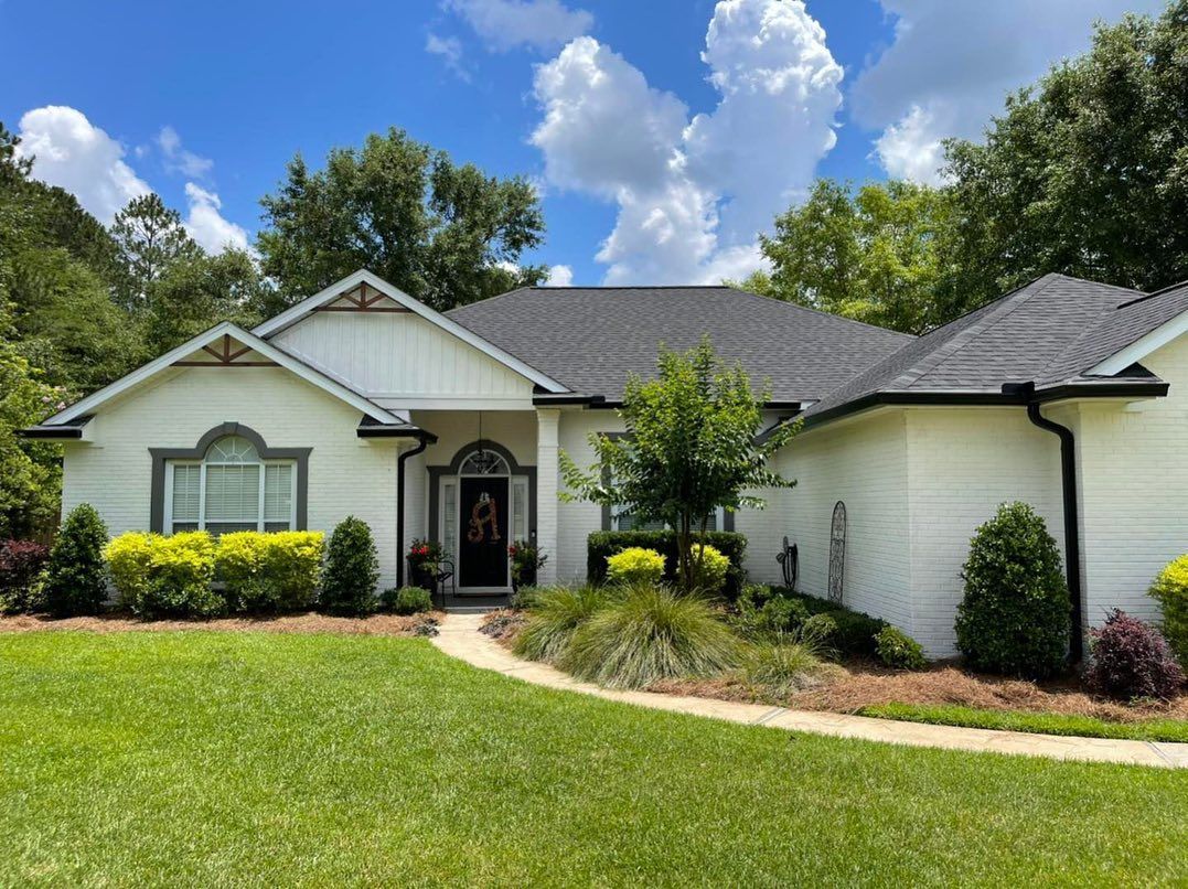 White brick house with a dark roof and black gutters on a sunny day. Green lawn and shrubs.