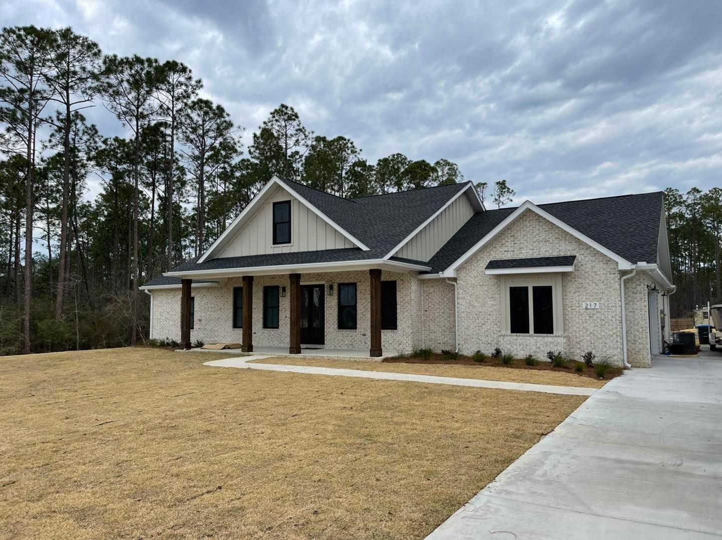 A brick house with a dark roof and wooden pillars sits on a grassy lot under a cloudy sky.