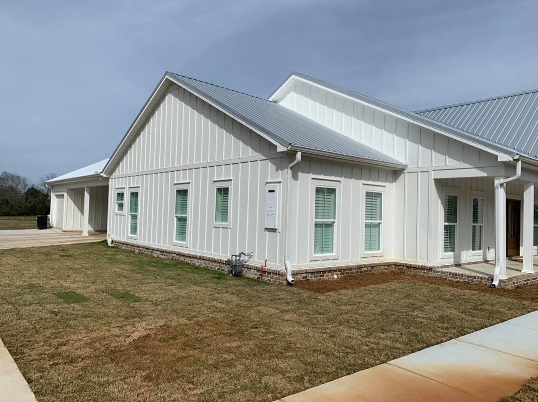 White farmhouse-style building with metal roof, windows, and green lawn. Cloudy sky.