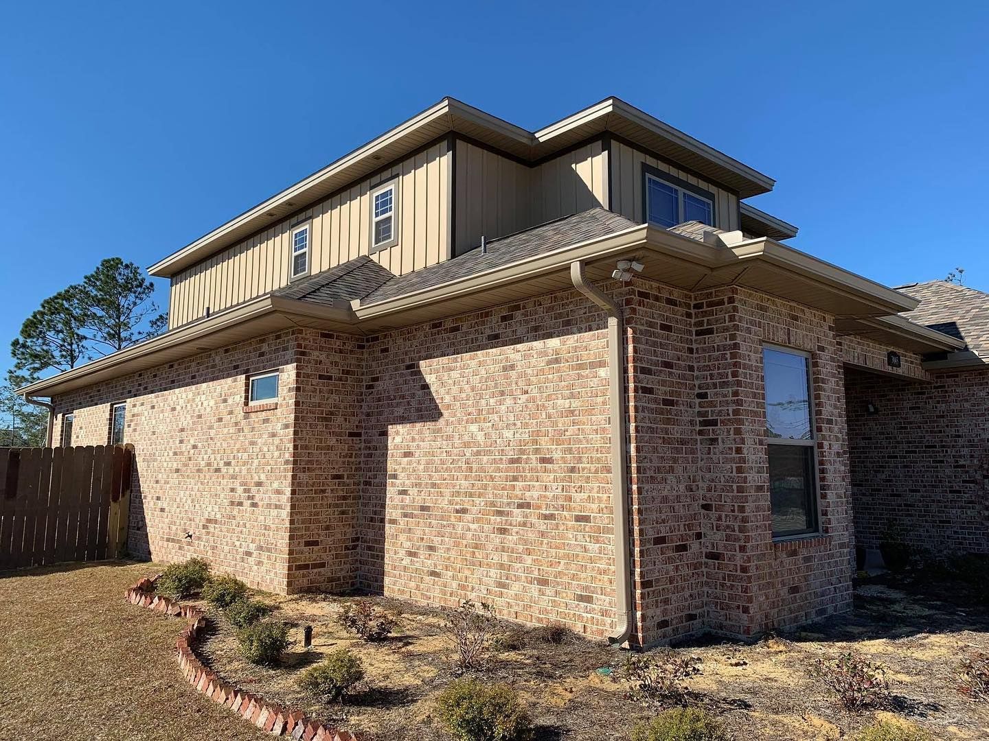 Two-story brick house with light brown siding, windows, and gutters against a blue sky.