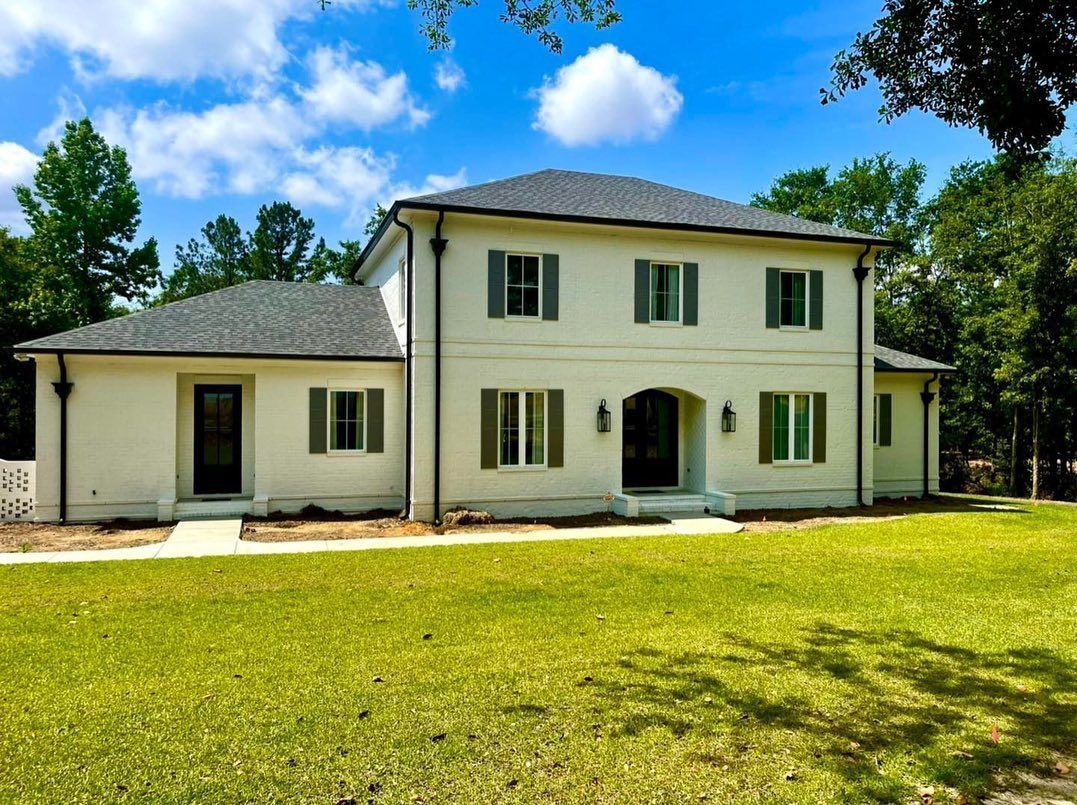Two-story white house with dark gray roof, green shutters, and green lawn on a sunny day.