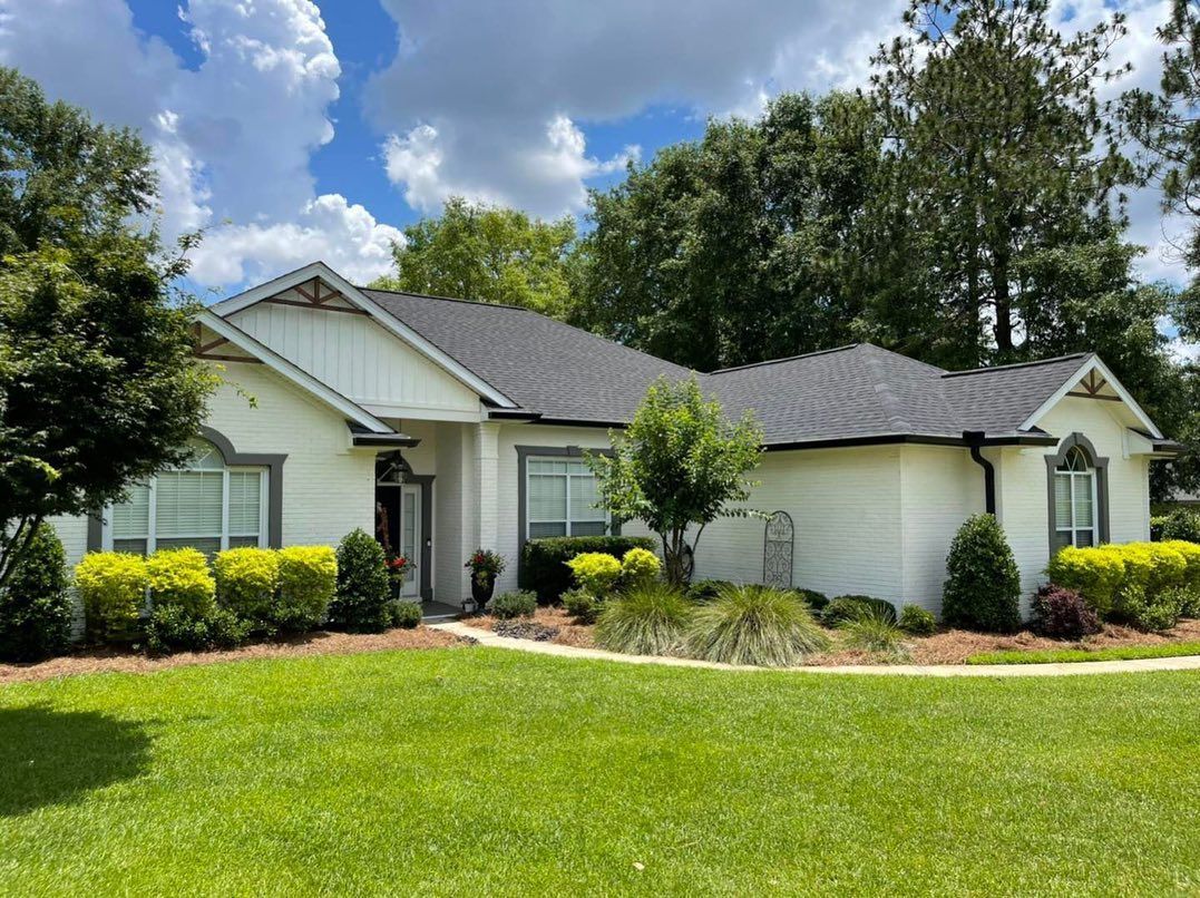 White house with black roof, green lawn, blue sky, and bushes.