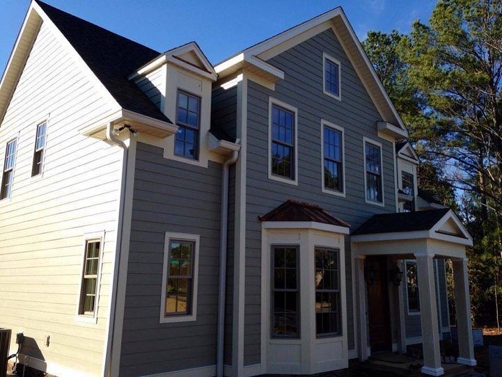 Two-story house with gray siding, white trim, and a small front porch, set in a sunny, wooded area.