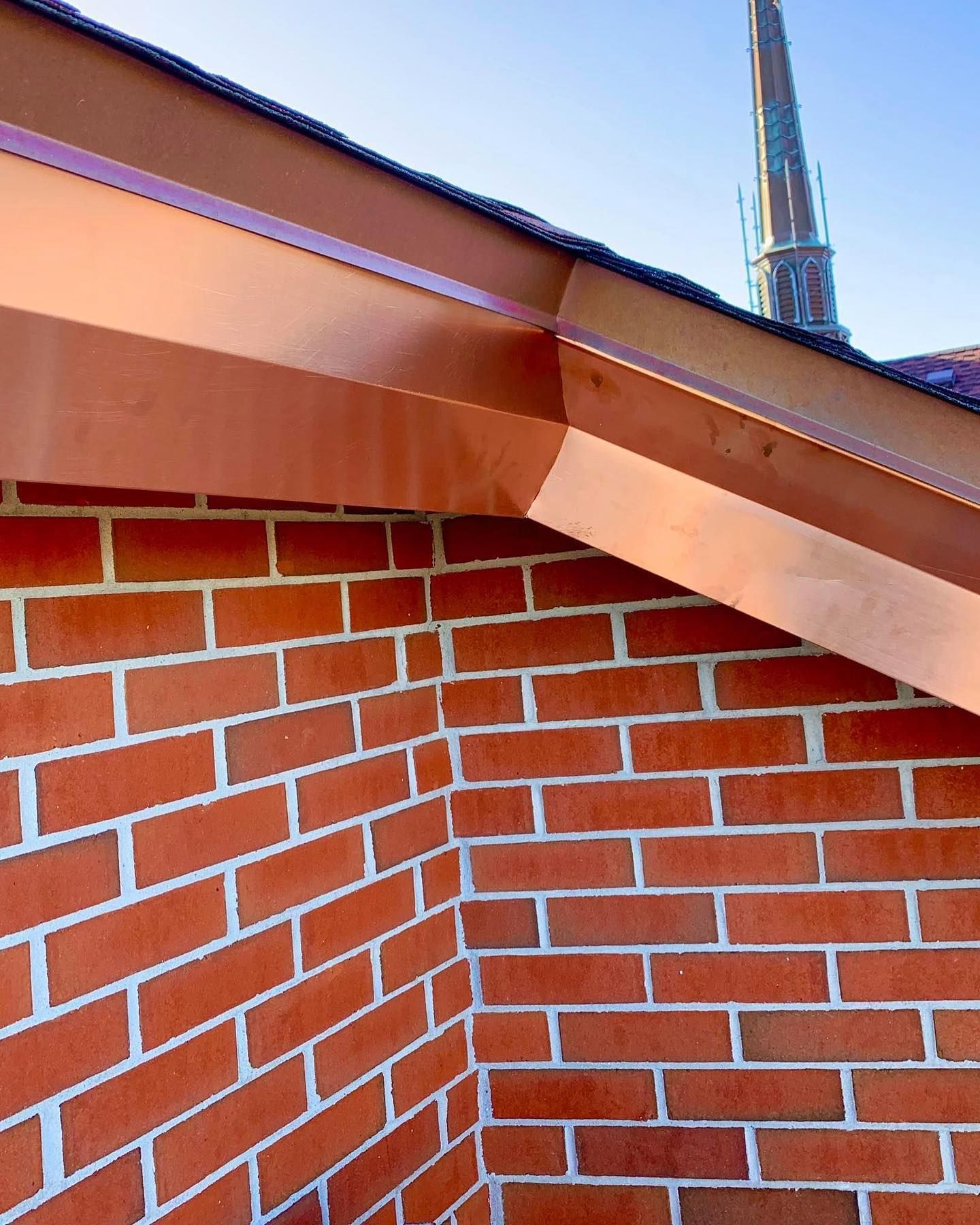 Copper-colored gutters on a brick building corner; church steeple visible in background.