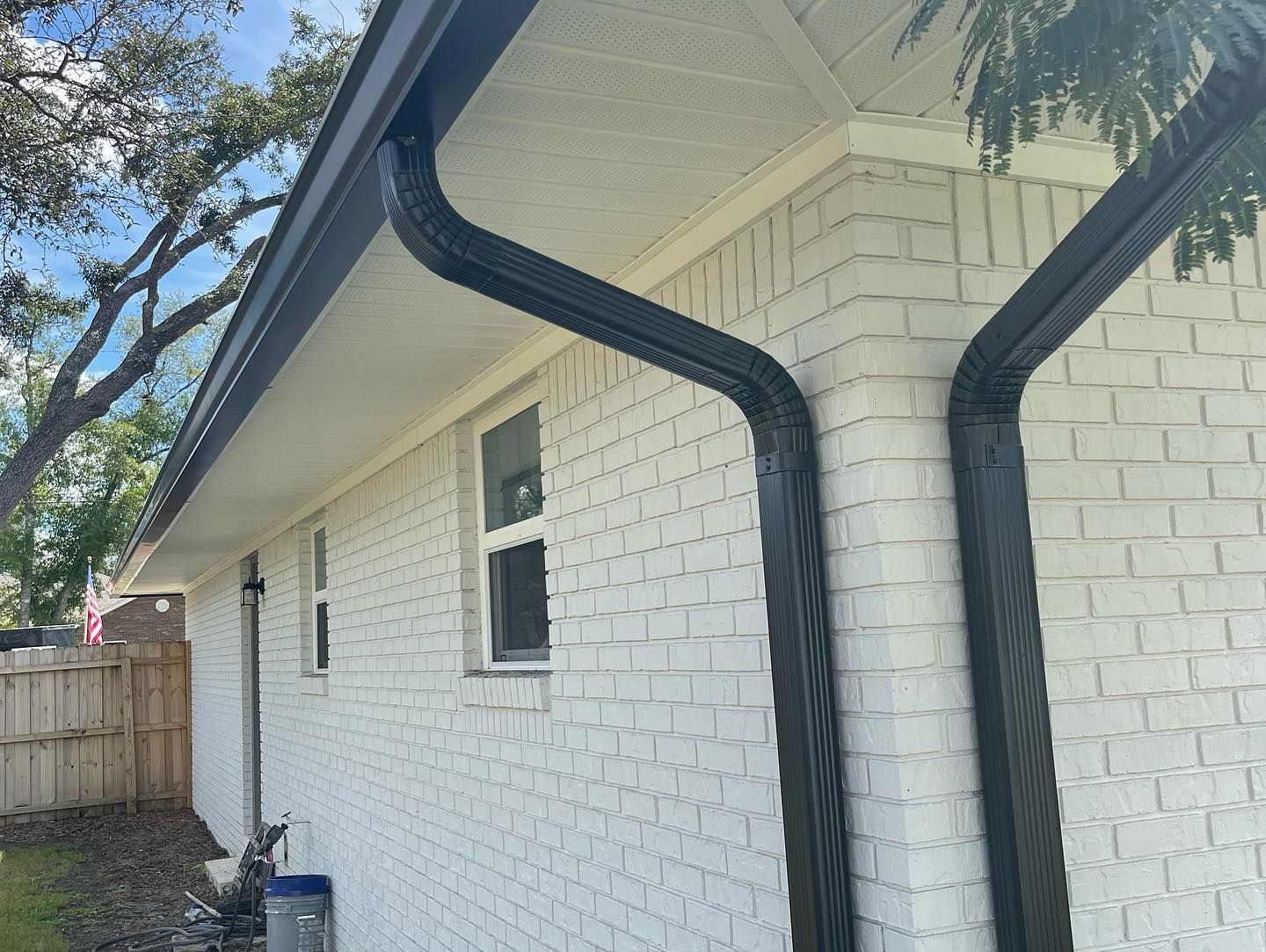 Black gutters on a white brick house, with a window and a wooden fence.