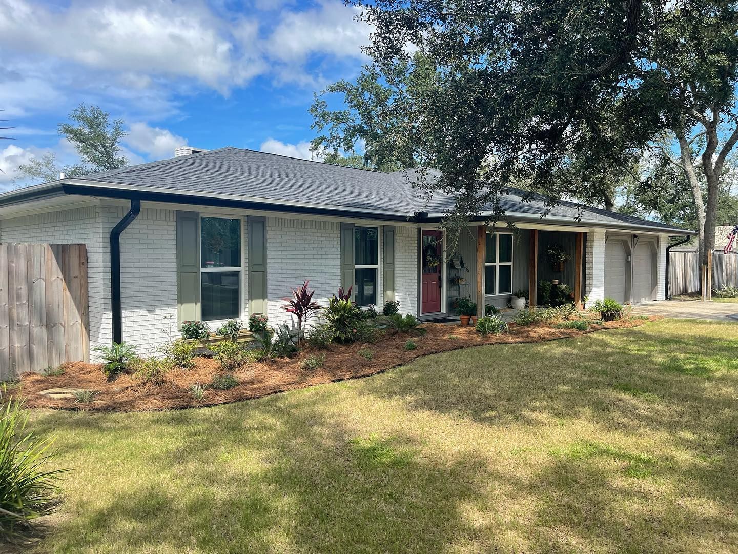 White-painted house with dark roof, shutters, and red door, with a grassy lawn and landscaped garden bed.