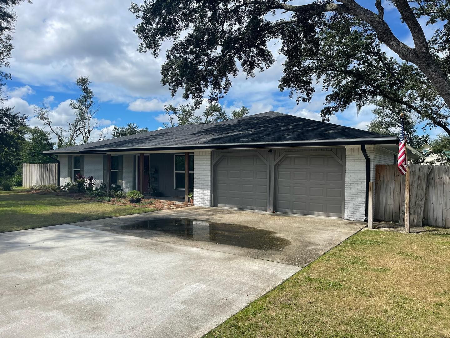 Gray house with two-car garage, large driveway with standing water, under a tree on a sunny day.