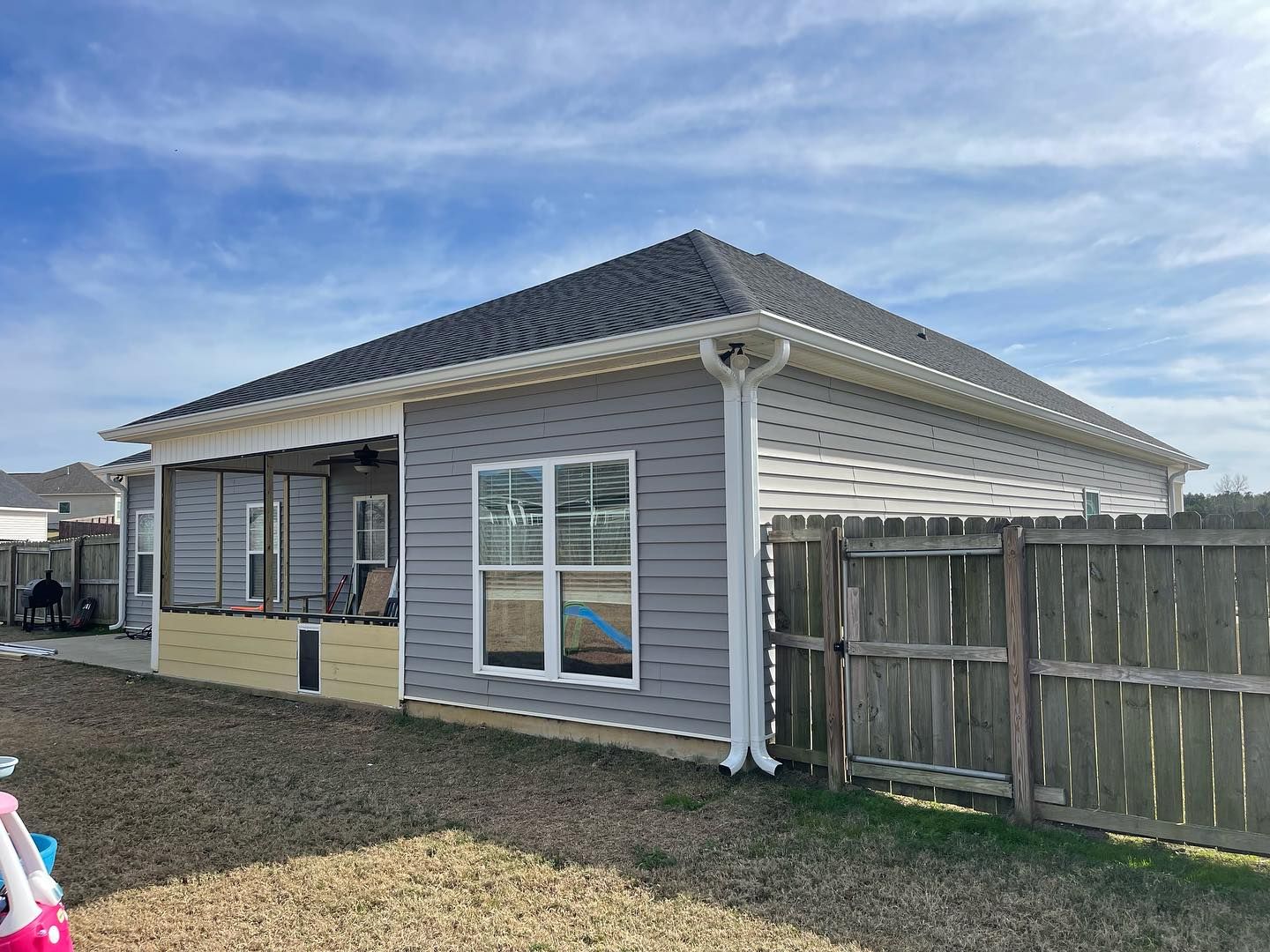 Gray house with a dark roof and white trim, next to a wooden fence under a blue sky.