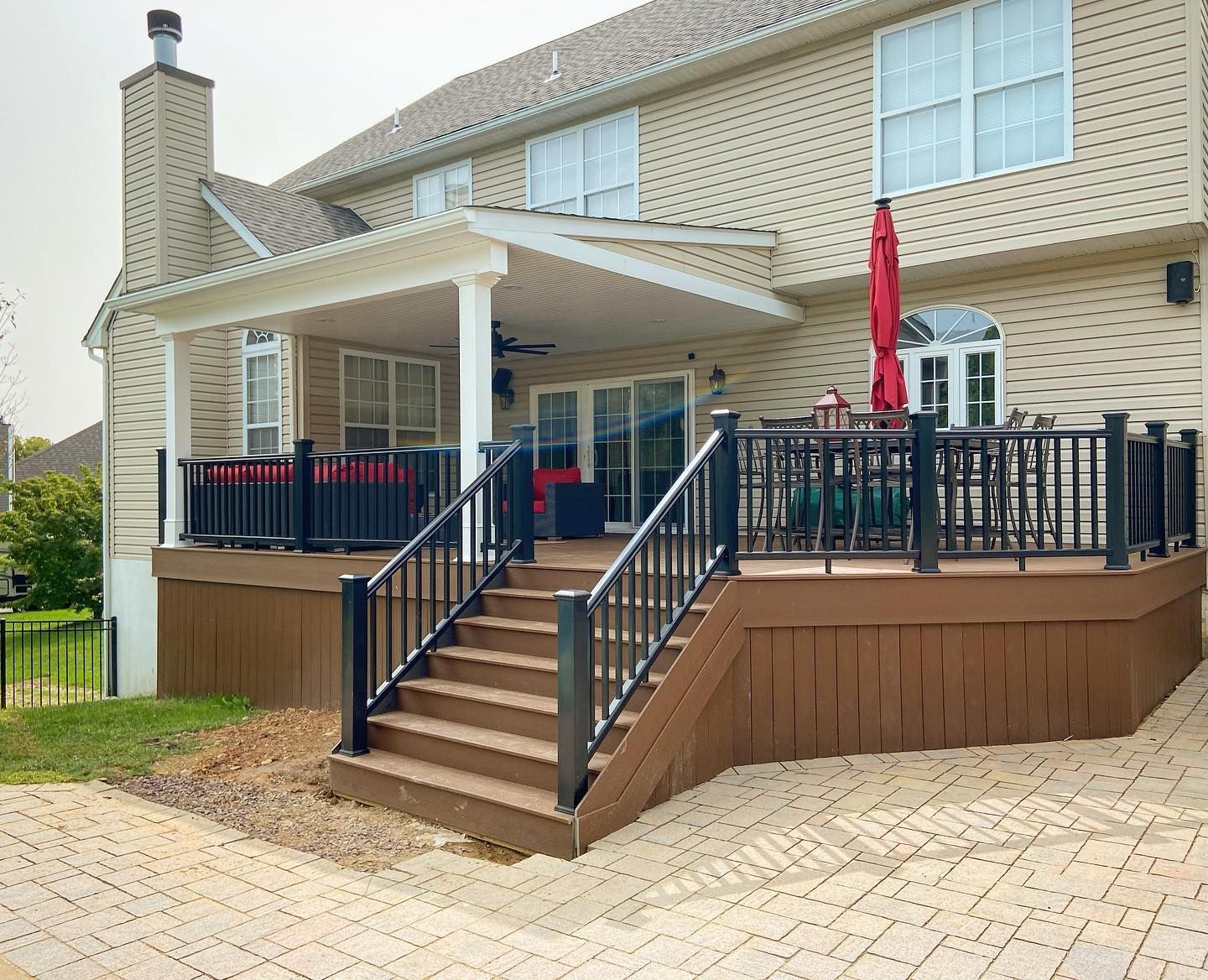 Backyard deck with stairs leading to a covered patio. Brown deck with black railing and seating. Beige siding on the house.