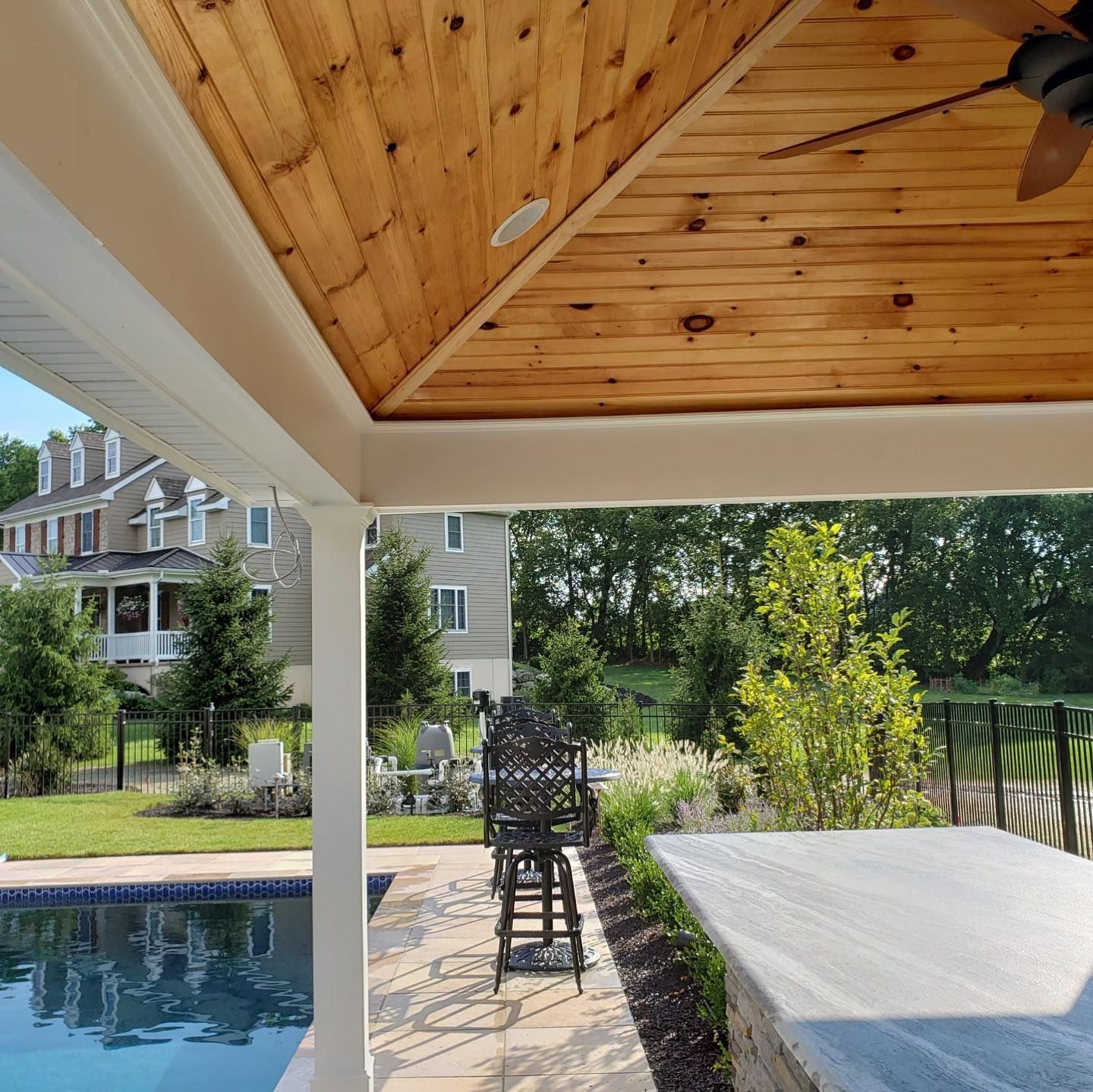 Patio with wooden ceiling and a pool in the foreground. Bar stools, trees, and houses in the background.