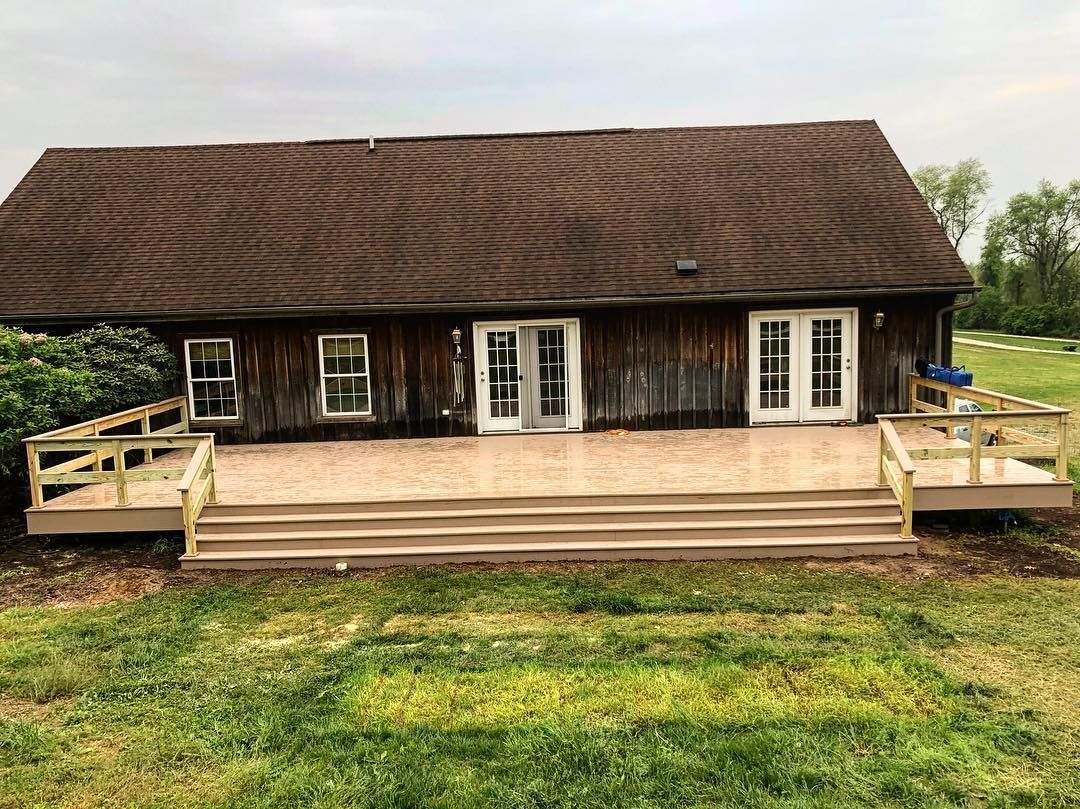 Back of a weathered brown building with a deck and steps, surrounded by green grass.