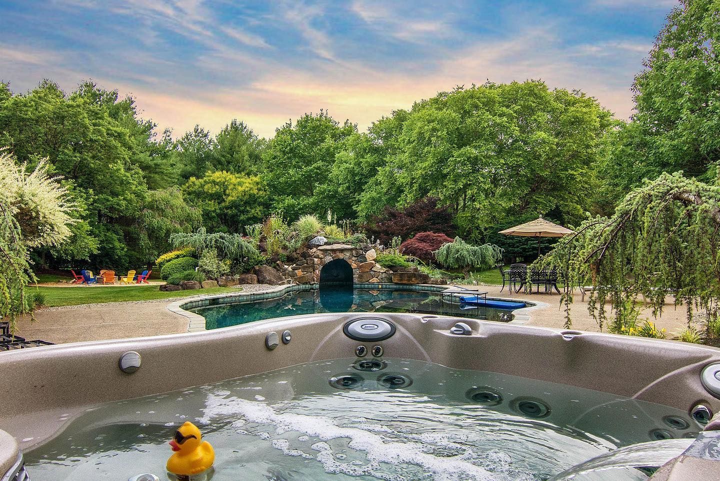 Hot tub with bubbly water, overlooking a pool and lush green backyard under a partly cloudy sky.