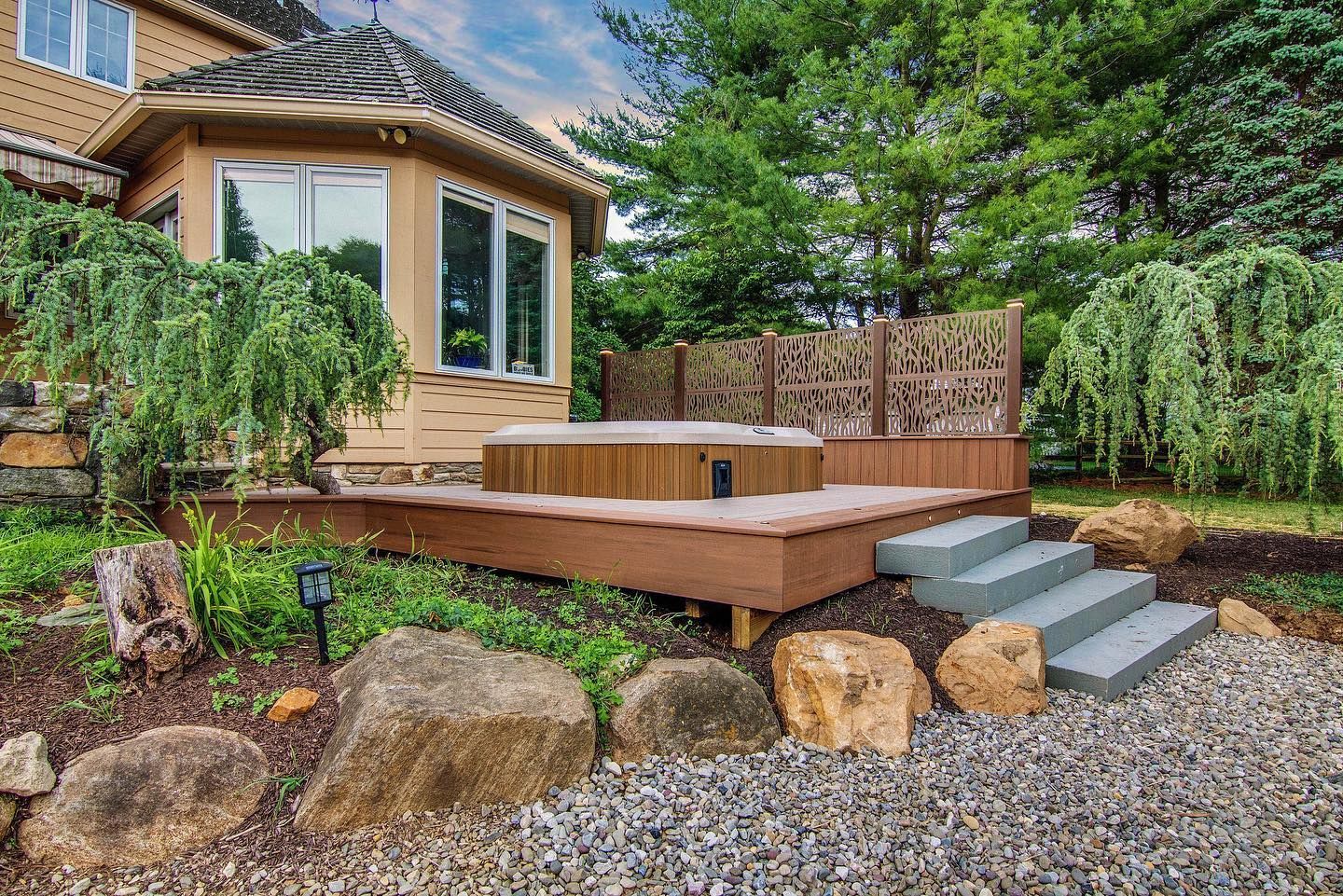 Wooden hot tub on a deck with steps, surrounded by rocks and trees.