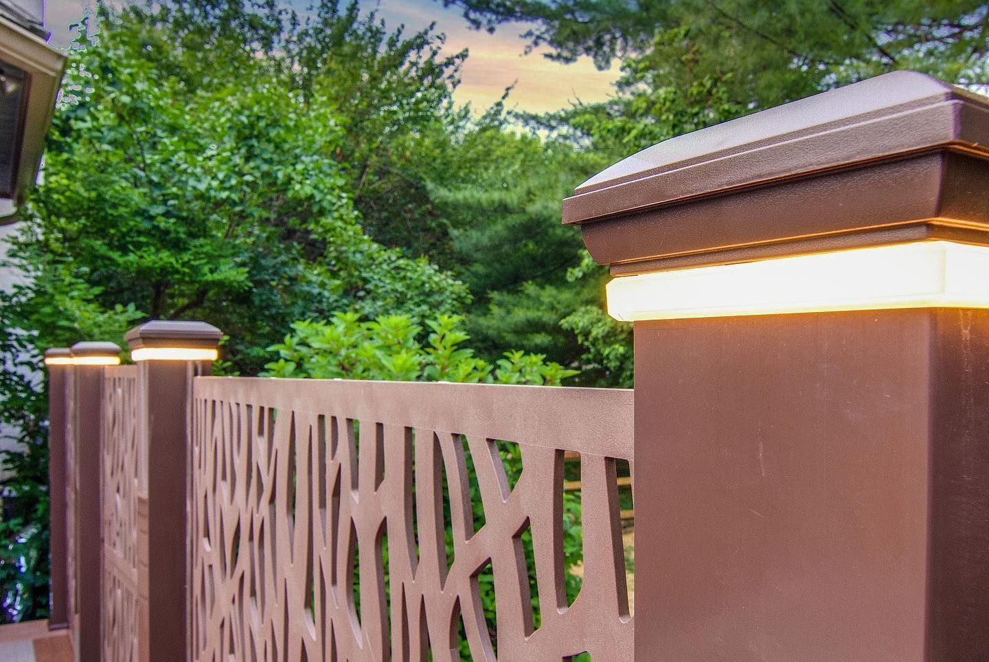 Brown metal fence with abstract cutouts, capped with lighted posts in a backyard with lush green trees.
