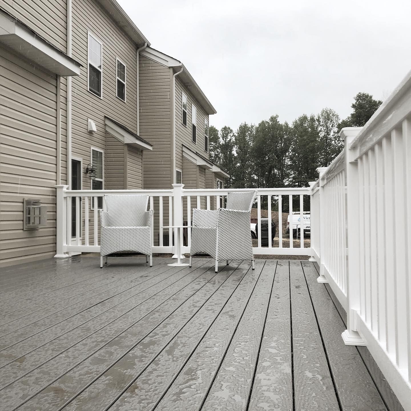Wooden deck with white railing and two chairs against a building with tan siding. Cloudy sky.