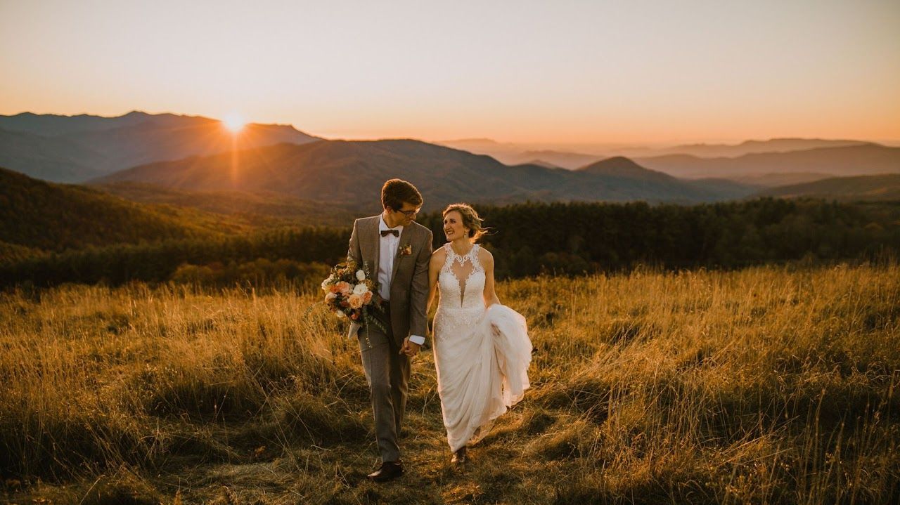 A bride and groom are walking through a field at sunset.