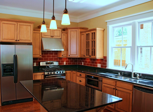 A modern kitchen with light wood cabinets, black granite countertops, a dark red tile backsplash, and stainless appliances.