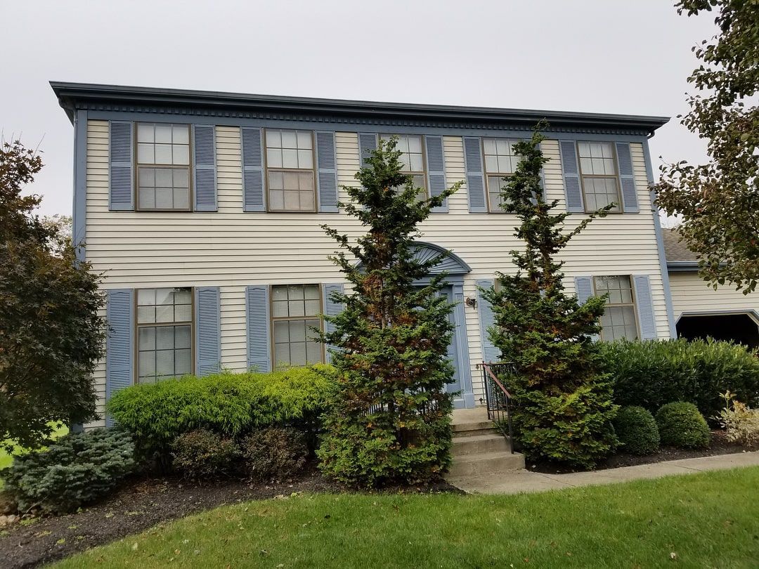 A two-story house with cream siding, blue shutters, and two tall, narrow evergreens flanking the front entrance.