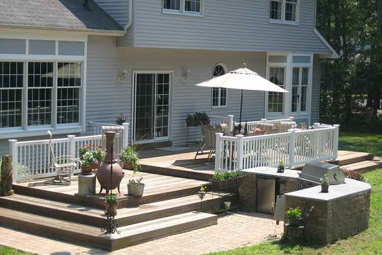 A multilevel wooden deck and patio with a dining set, white railings, and an outdoor kitchen area behind a suburban home.