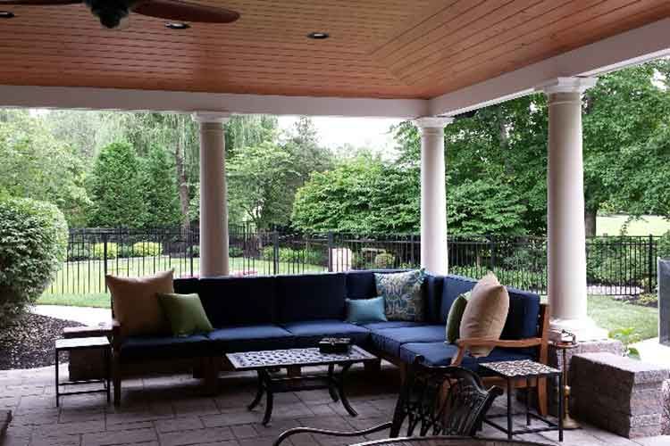 A covered outdoor patio featuring a large navy blue sectional sofa, a coffee table, and lush greenery in the background.