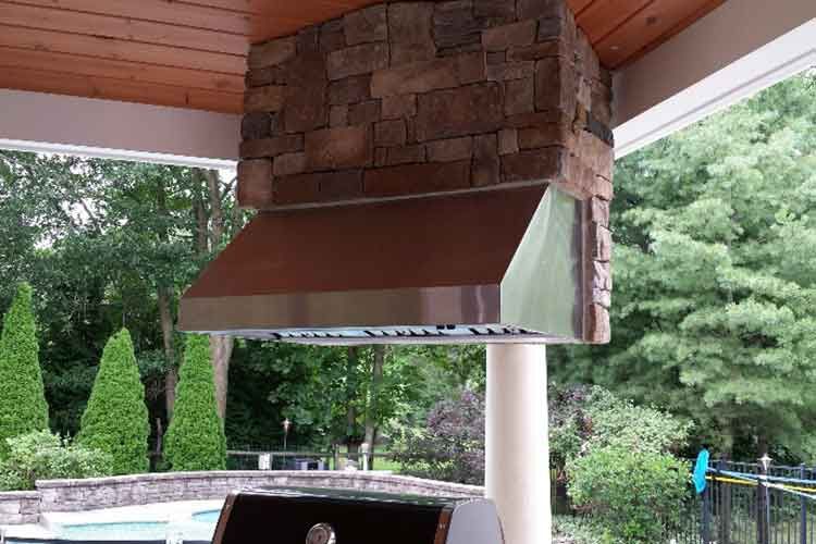 A copper outdoor range hood installed beneath a wood-paneled patio ceiling, with a stone chimney stack behind it.