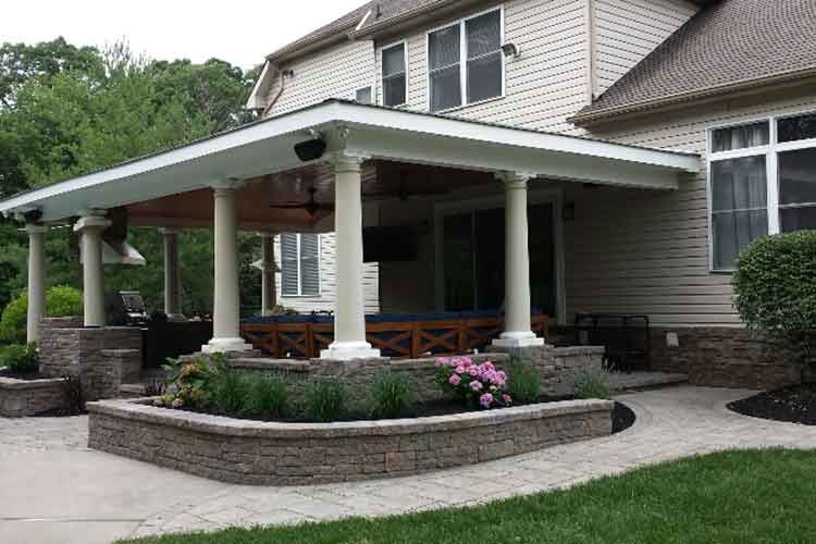 A covered patio area attached to a beige house, featuring white columns, stone wall planters, and a brick paver walkway.