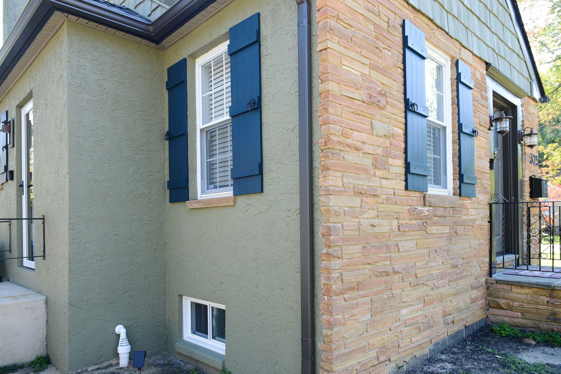 Exterior corner of a house featuring tan stone siding, olive green stucco, dark blue window shutters, and a front porch.