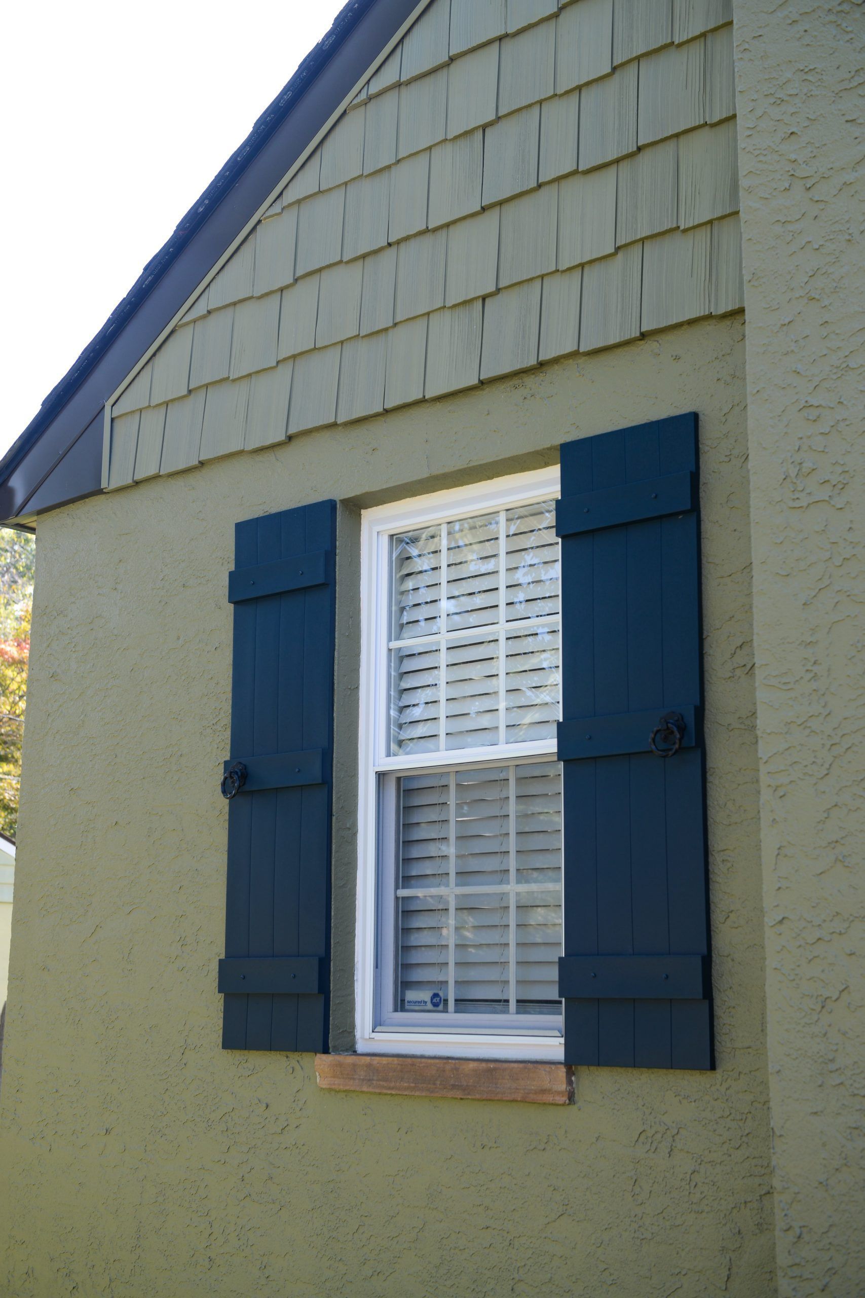 A sage green house exterior featuring a window with dark blue shutters and cedar shake siding in the gable.