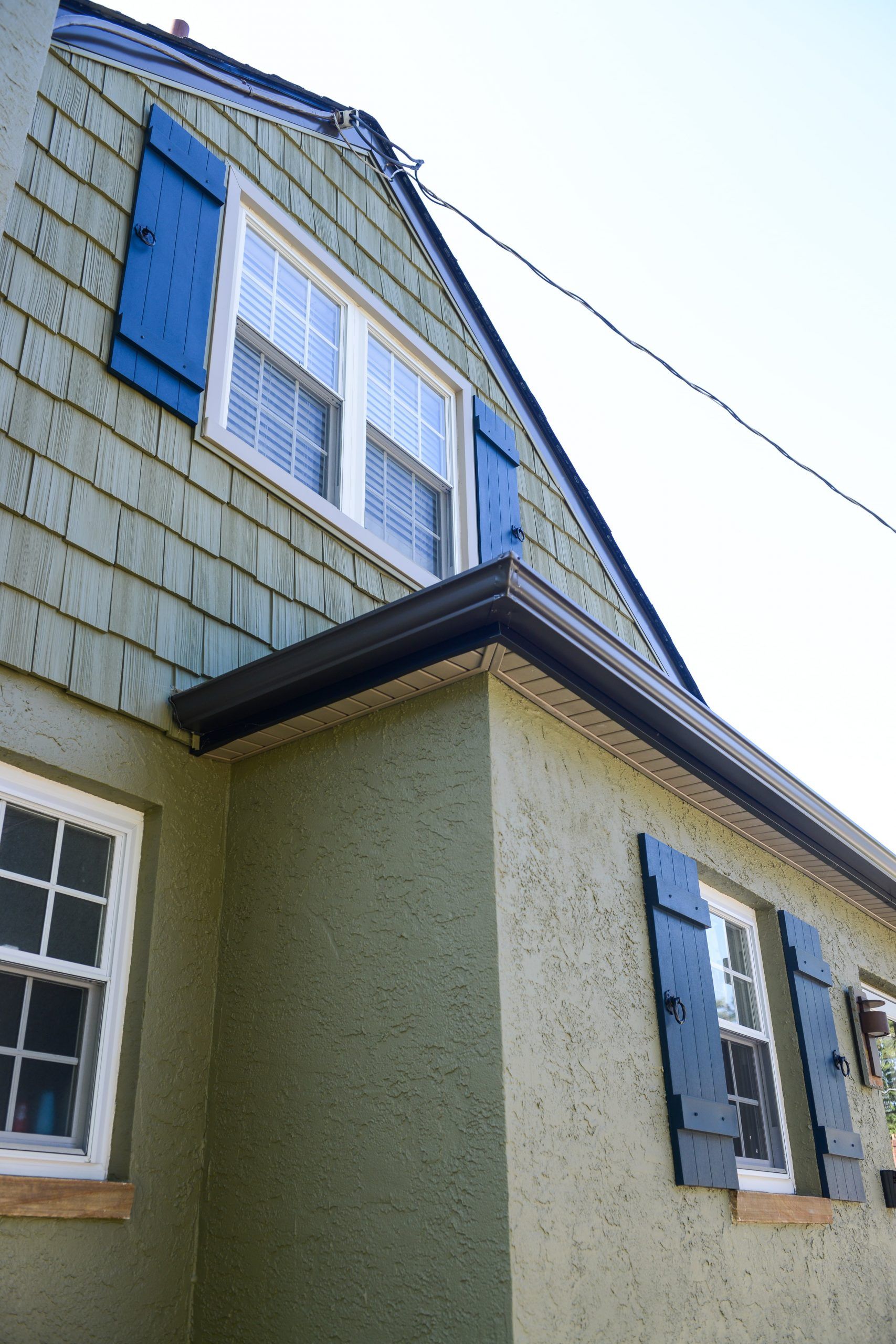 A low-angle view of a house exterior with green stucco siding, blue shutters on the windows, and a dark brown gutter.