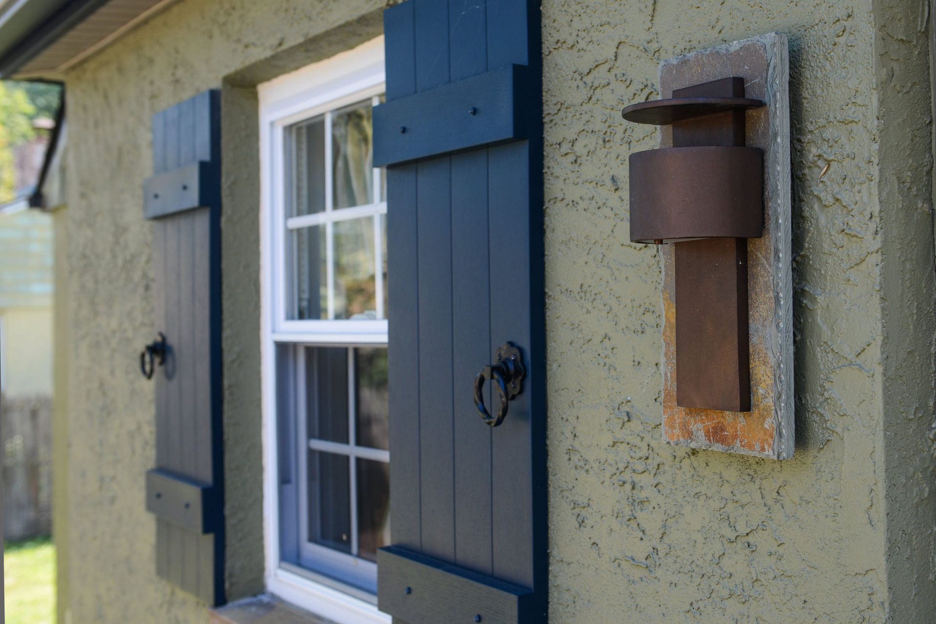 Exterior view of a dark window with black shutters and a brown wall-mounted light fixture on textured green stucco.