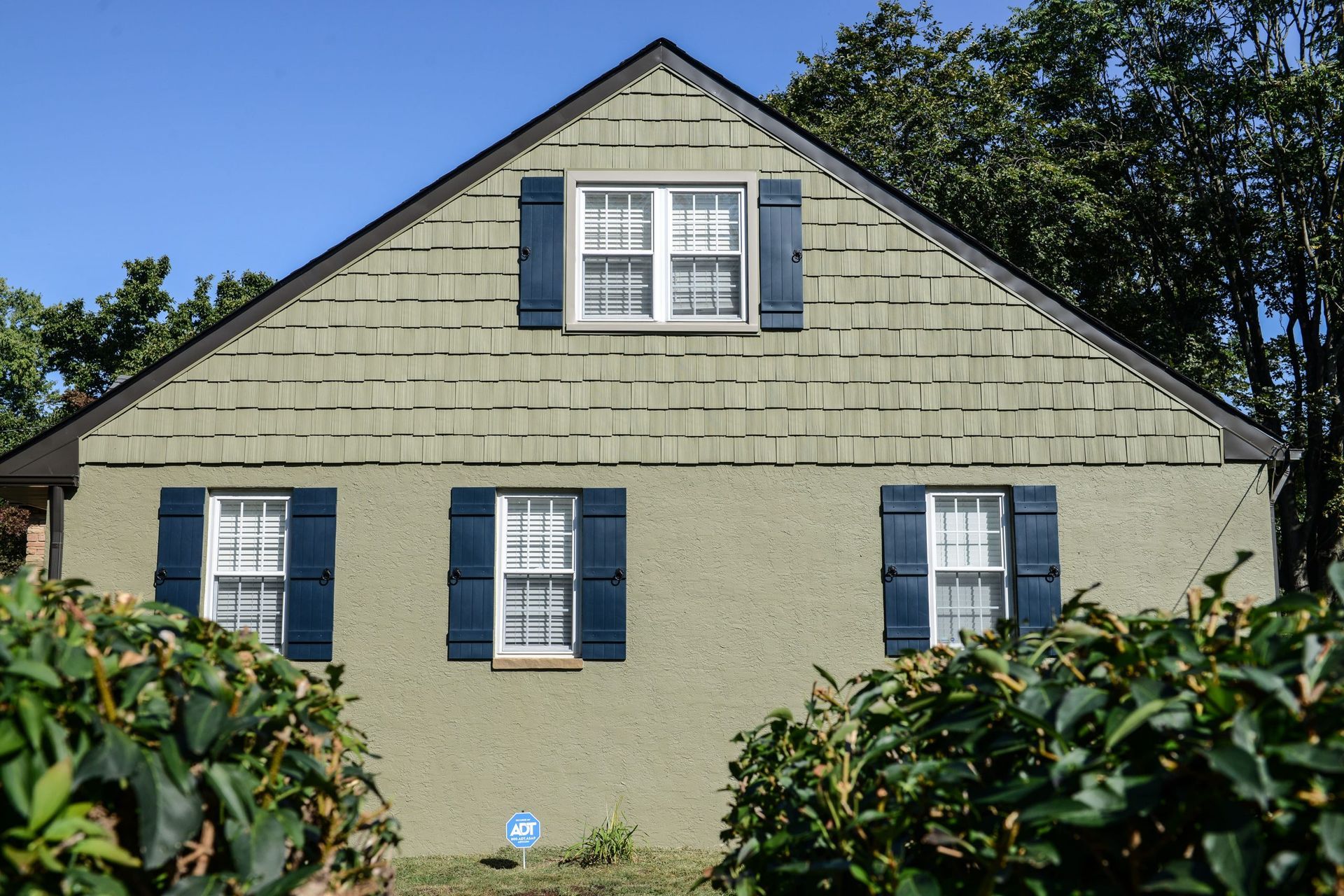 A two-story, sage-green house facade with textured shingle siding above, stucco below, and dark blue window shutters.