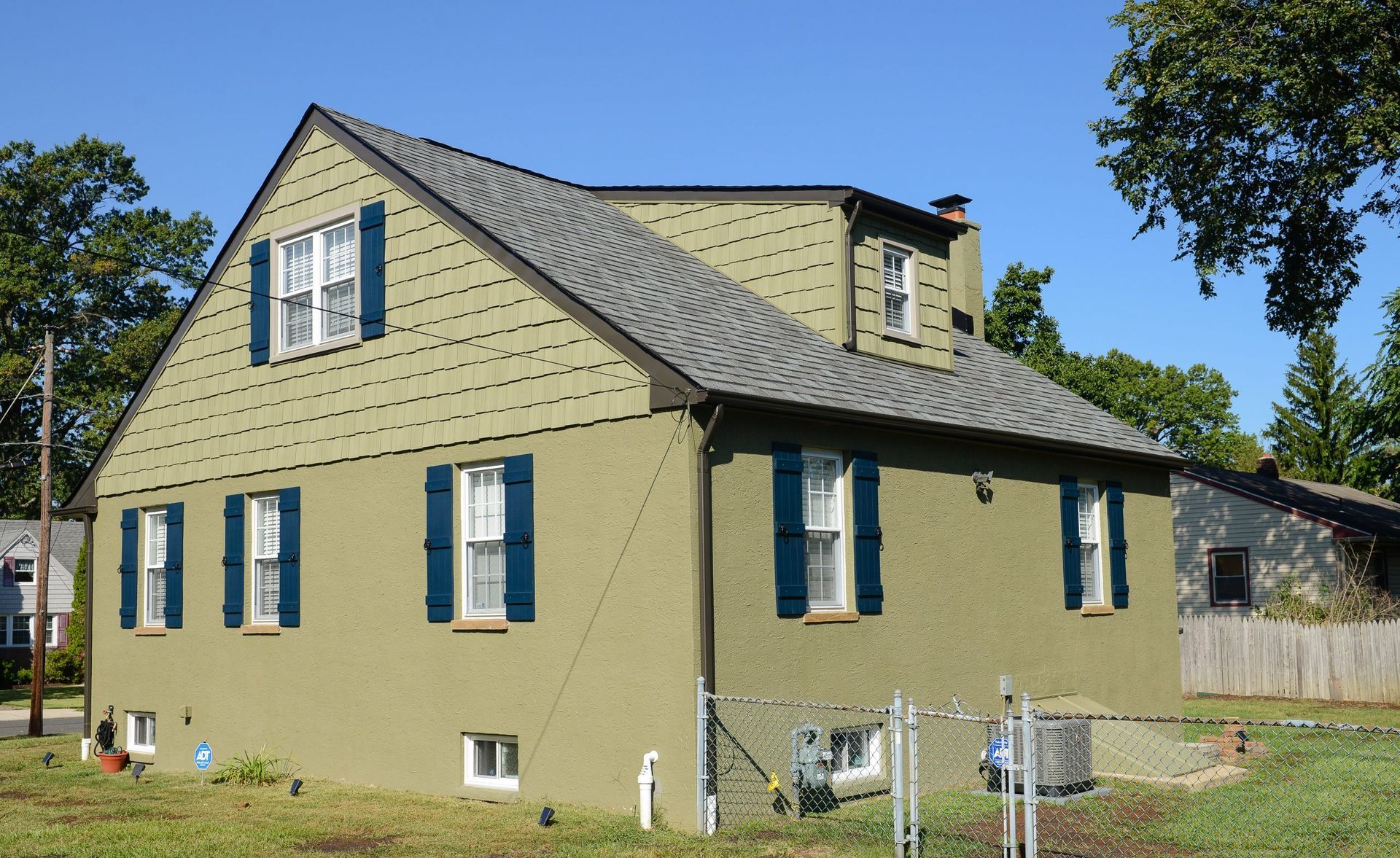 A two-story green stucco house with a shingled roof, dark blue shutters, and a dormer window on a sunny day.