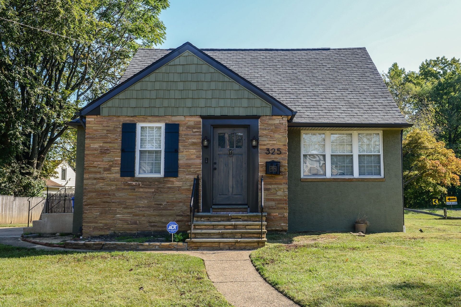 A small, one-story house with stone and olive-green siding, a dark roof, a front door, and a brick path on a grassy lawn.