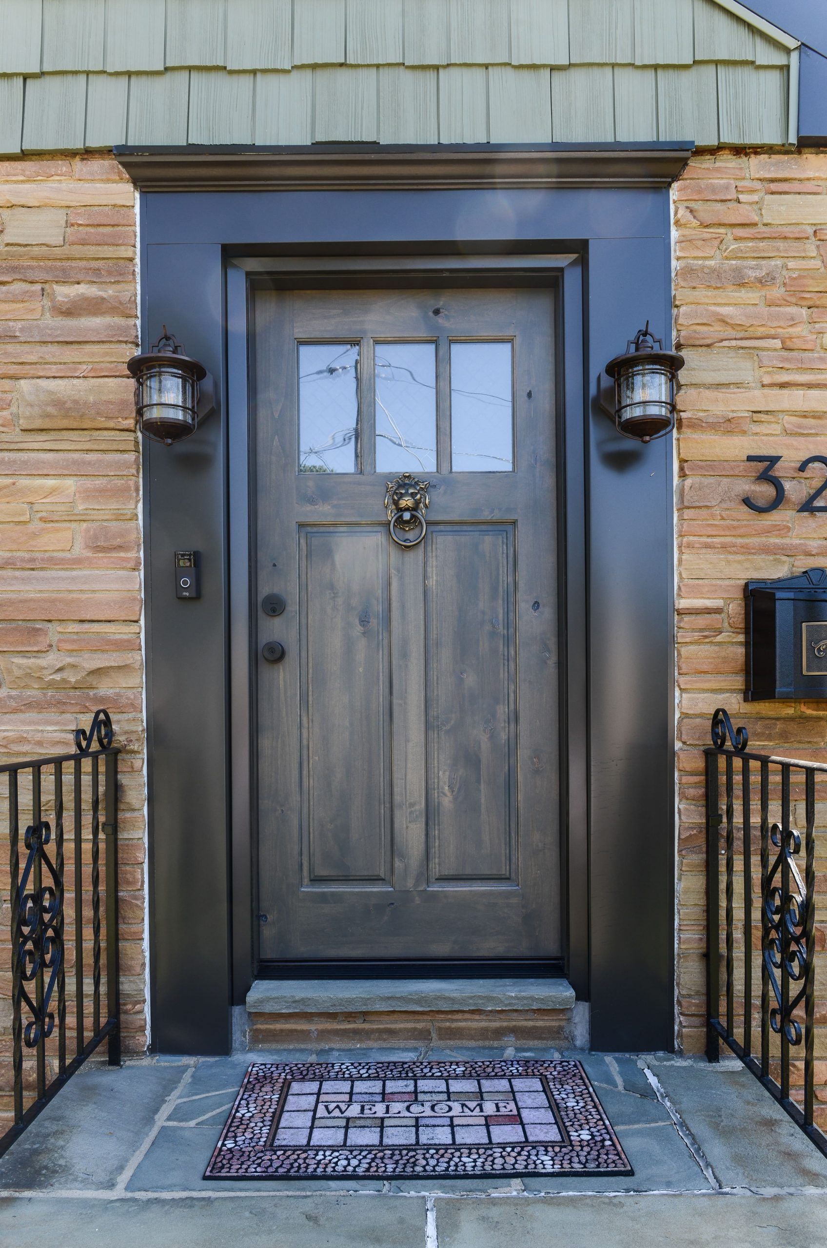 A dark brown wooden front door with a lion door knocker and glass panels, set in a stone wall with black iron railings.