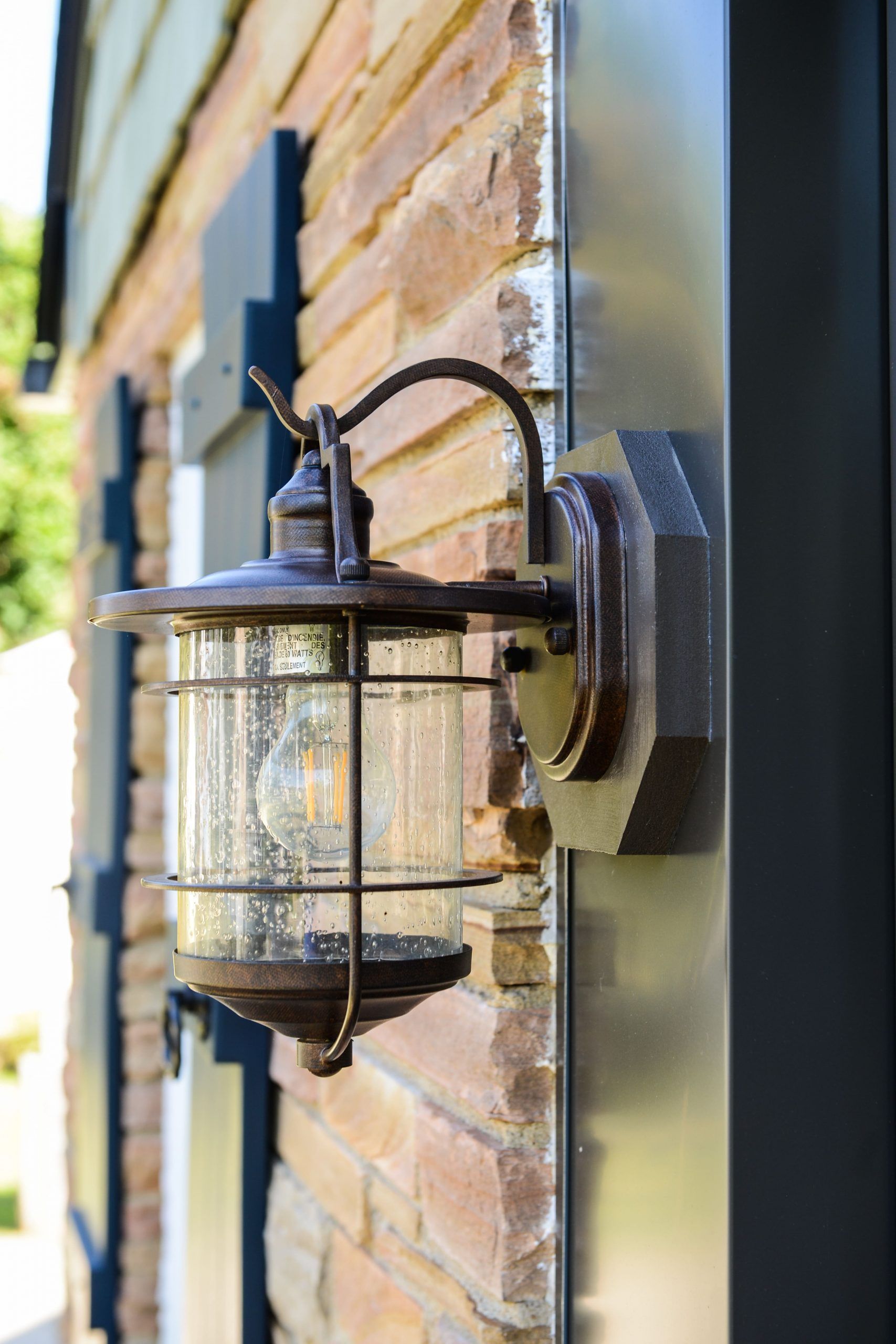 A vintage-style outdoor wall lantern with a seeded glass shade mounted on a brick wall next to dark window shutters.