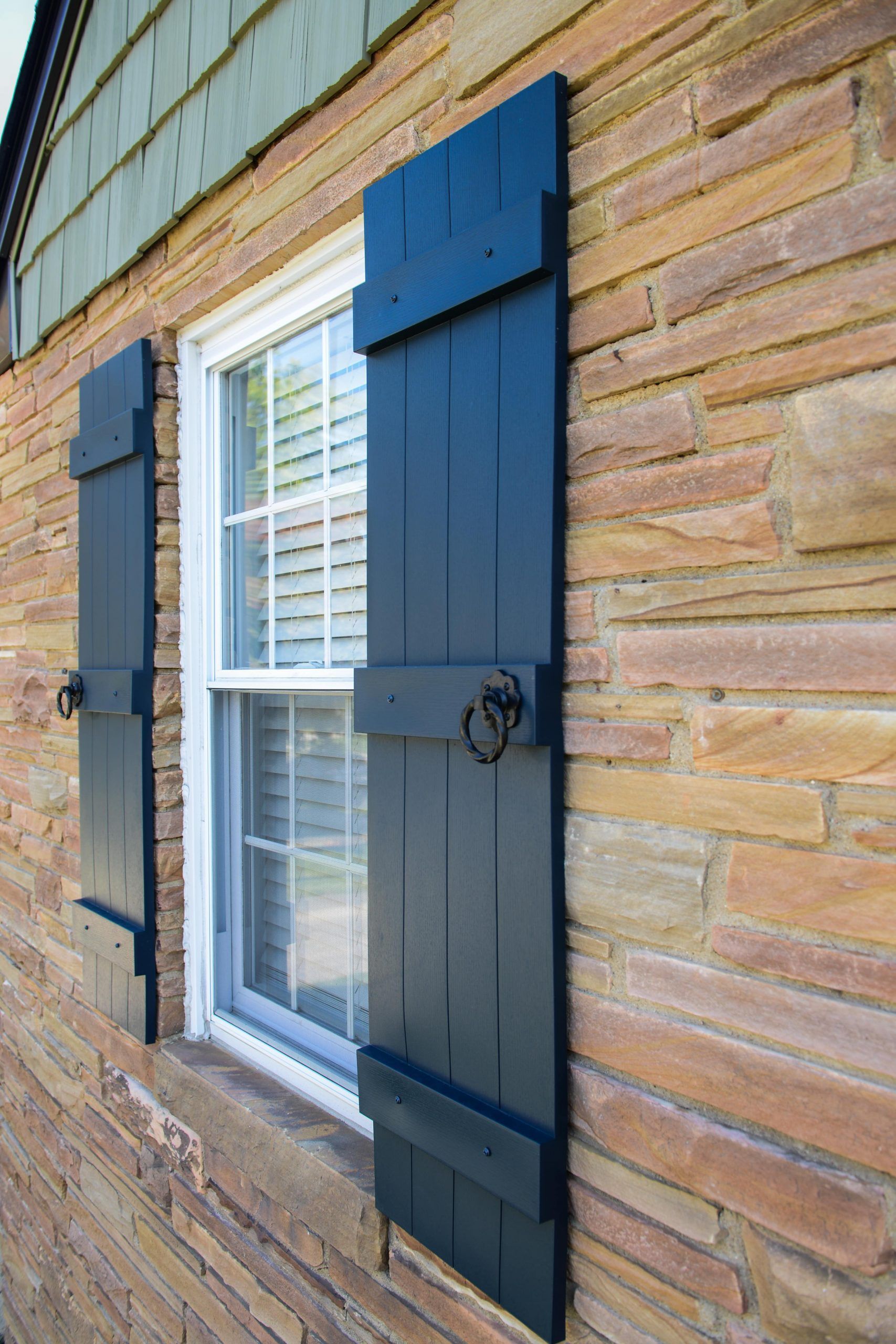 Black rustic shutters with decorative ring hardware flanking a window on a textured stone exterior wall.