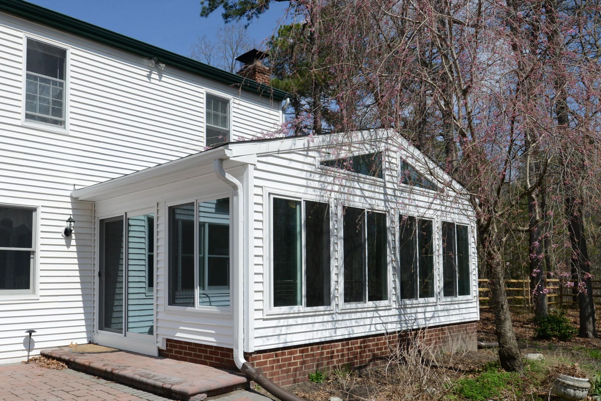 A white, multi-windowed sunroom addition attached to the side of a white siding house, with a brick foundation base.