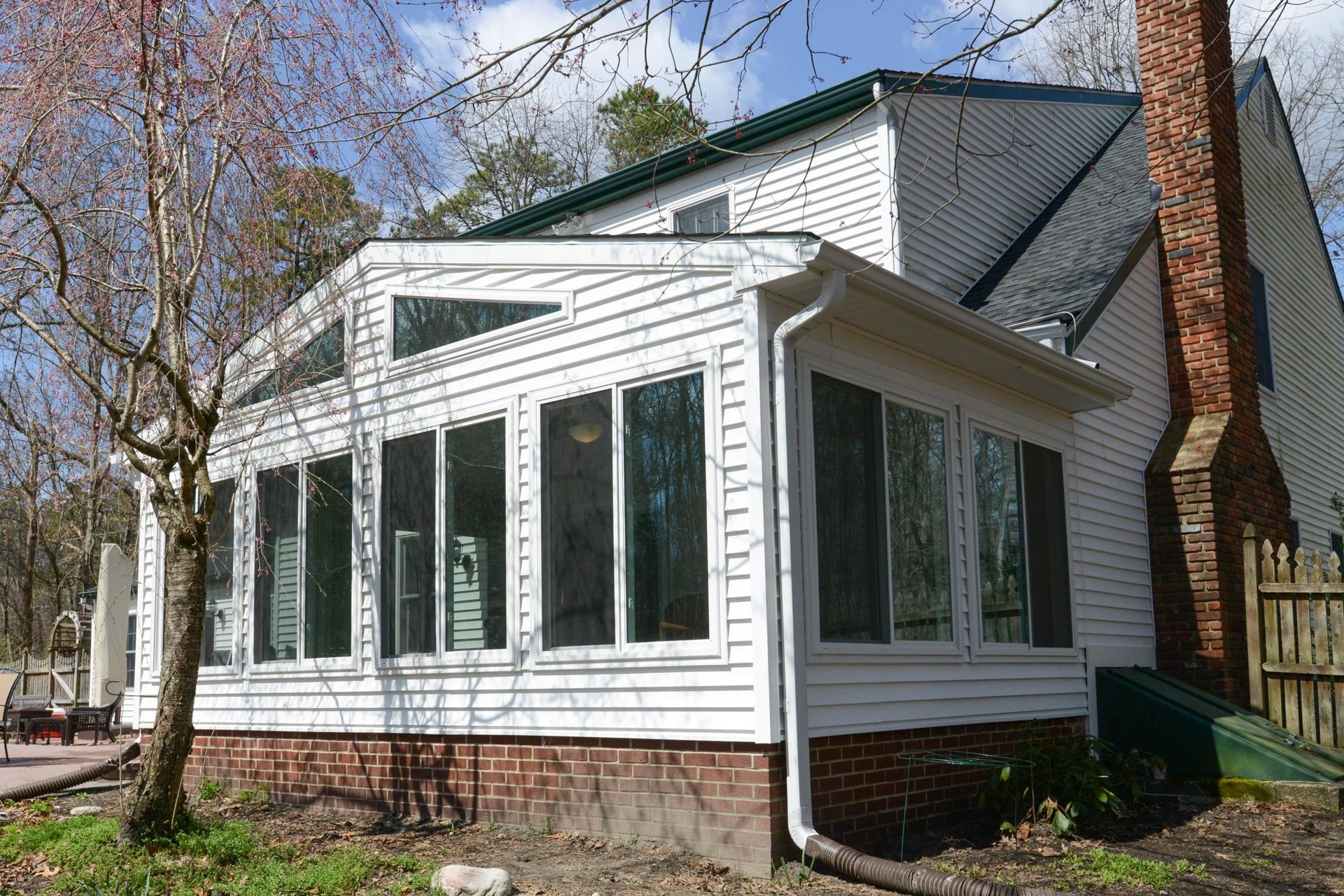A white sunroom addition with glass windows attached to the side of a house with a brick chimney and white siding.