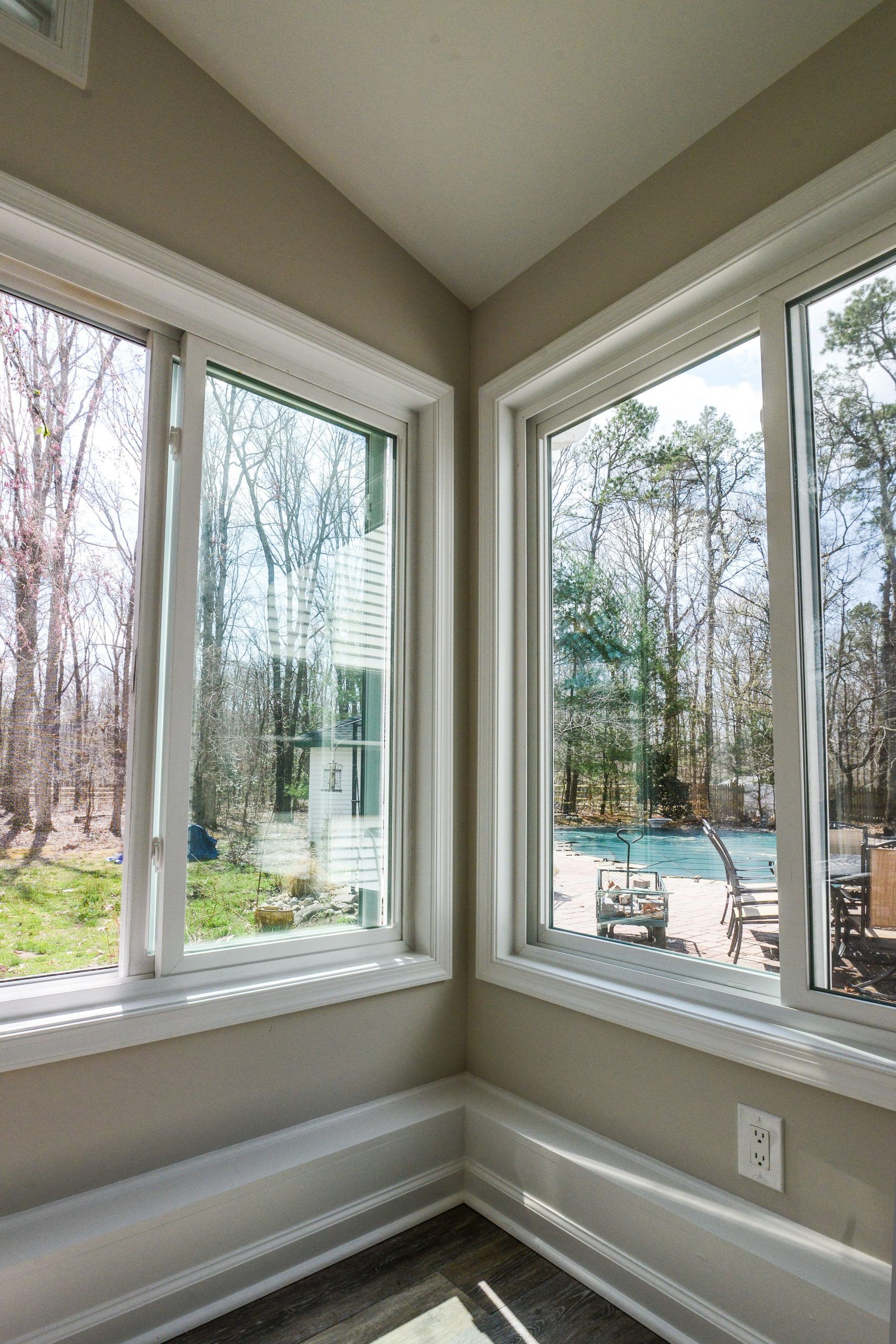 Corner view of two large windows looking out onto a sunny backyard with trees, green lawn, and a swimming pool.