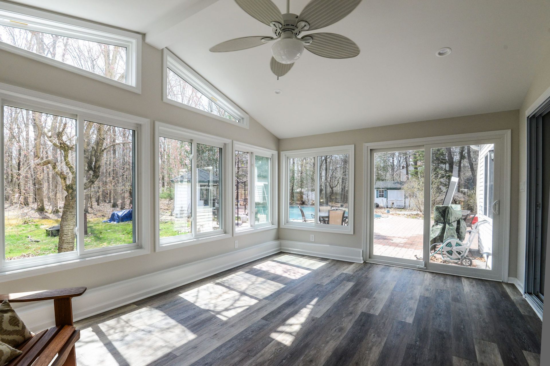 A sunroom with dark wood-look floors, a ceiling fan, and large windows looking out onto a wooded yard.