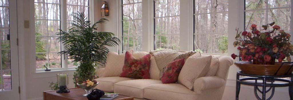 A sunroom with a beige sofa, two patterned throw pillows, a coffee table with a plant, and a potted arrangement nearby.