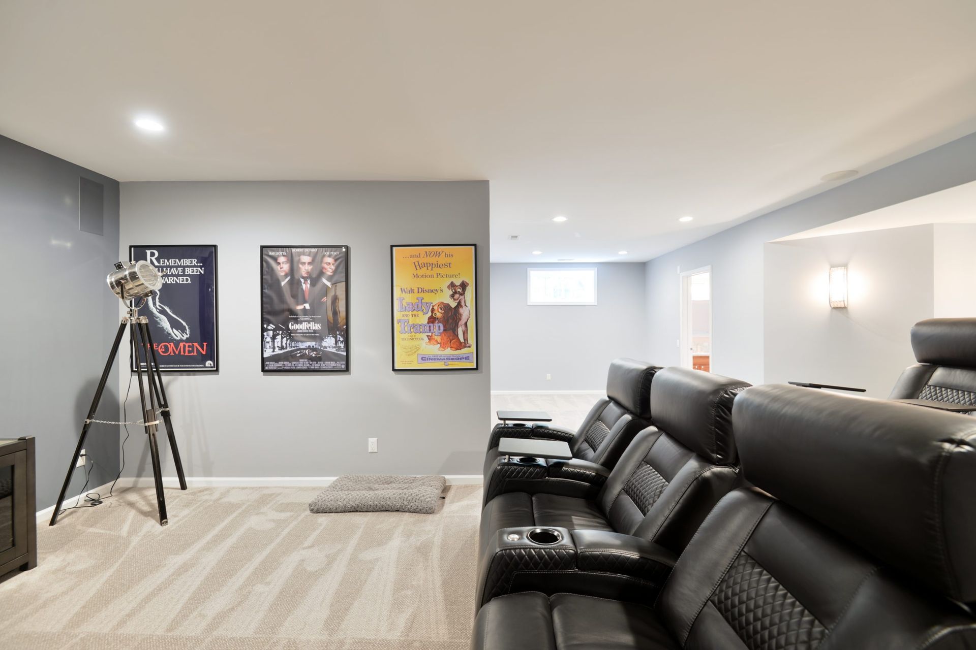 A home theater room with black reclining seats, gray walls, a tripod floor lamp, and three movie posters.