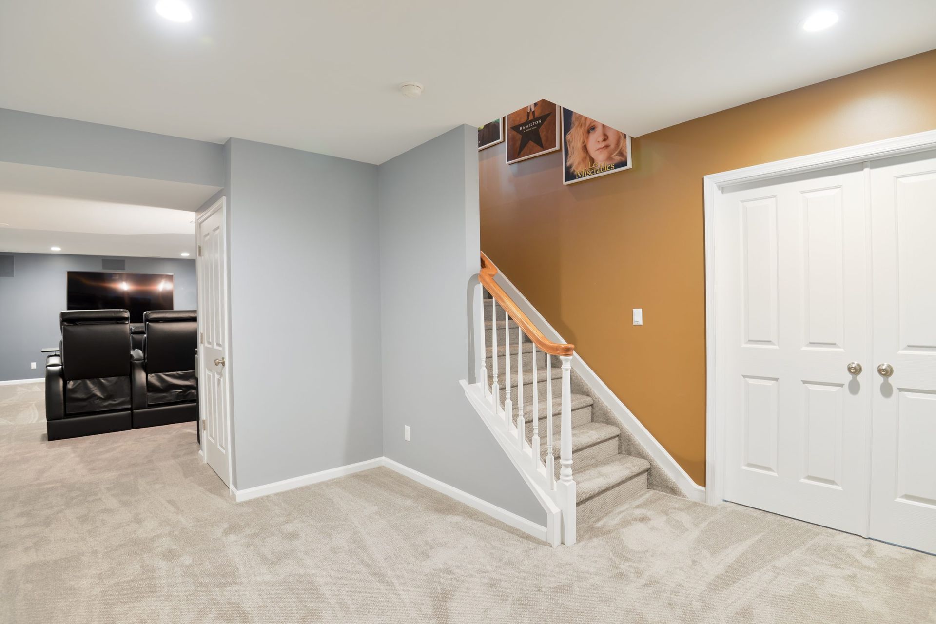 A basement with gray carpet, a gray accent wall, stairs with a white railing, a brown wall, and double white doors.