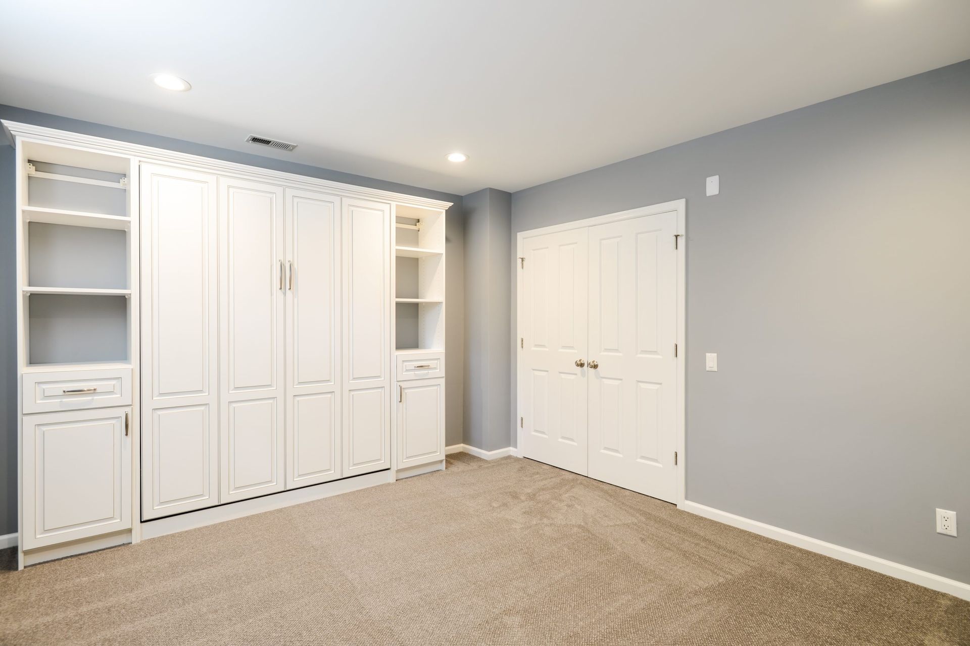 A room with beige carpet, light gray walls, a white built-in wall unit with shelves, and a closed white double door.