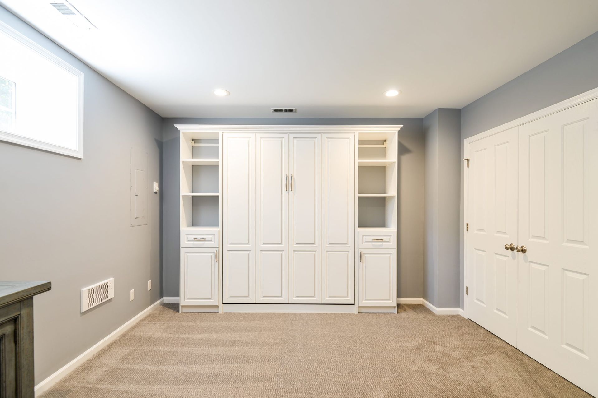 A white Murphy bed with built-in shelving centered on a light gray wall in a carpeted room with double closet doors.