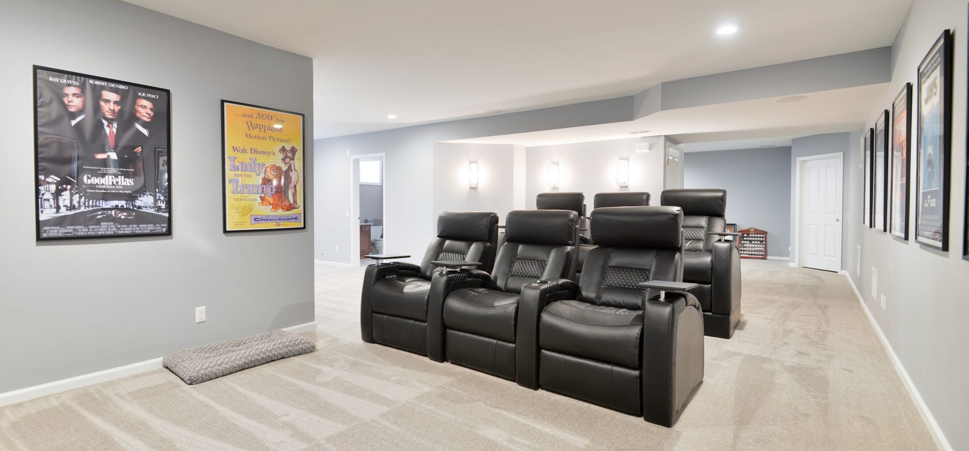 A home theater room with three black leather reclining seats, grey carpet, and framed movie posters on the walls.
