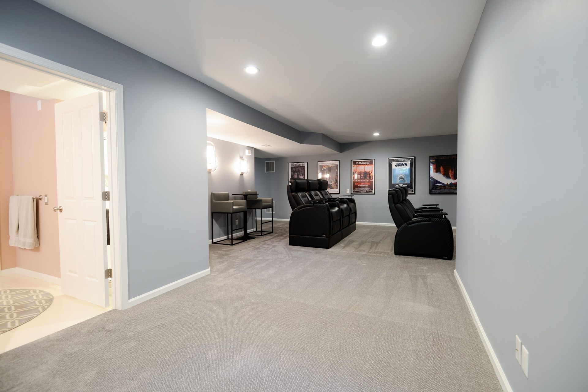 A carpeted basement home theater with two rows of black leather recliners and framed movie posters on the walls.