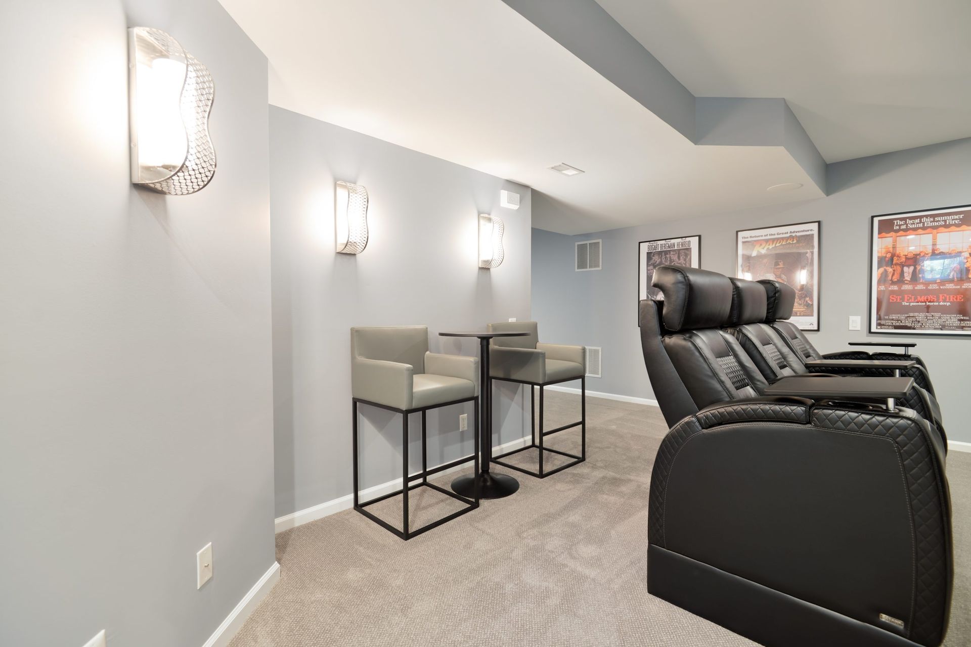 Home theater room with grey walls, a small table with two bar stools, and a row of black reclining cinema chairs.